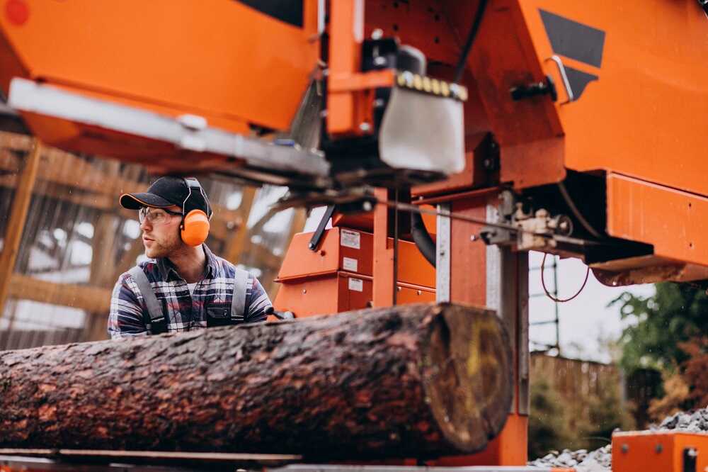 Tree service worker in protective gear operating a large orange sawmill machine cutting a log.