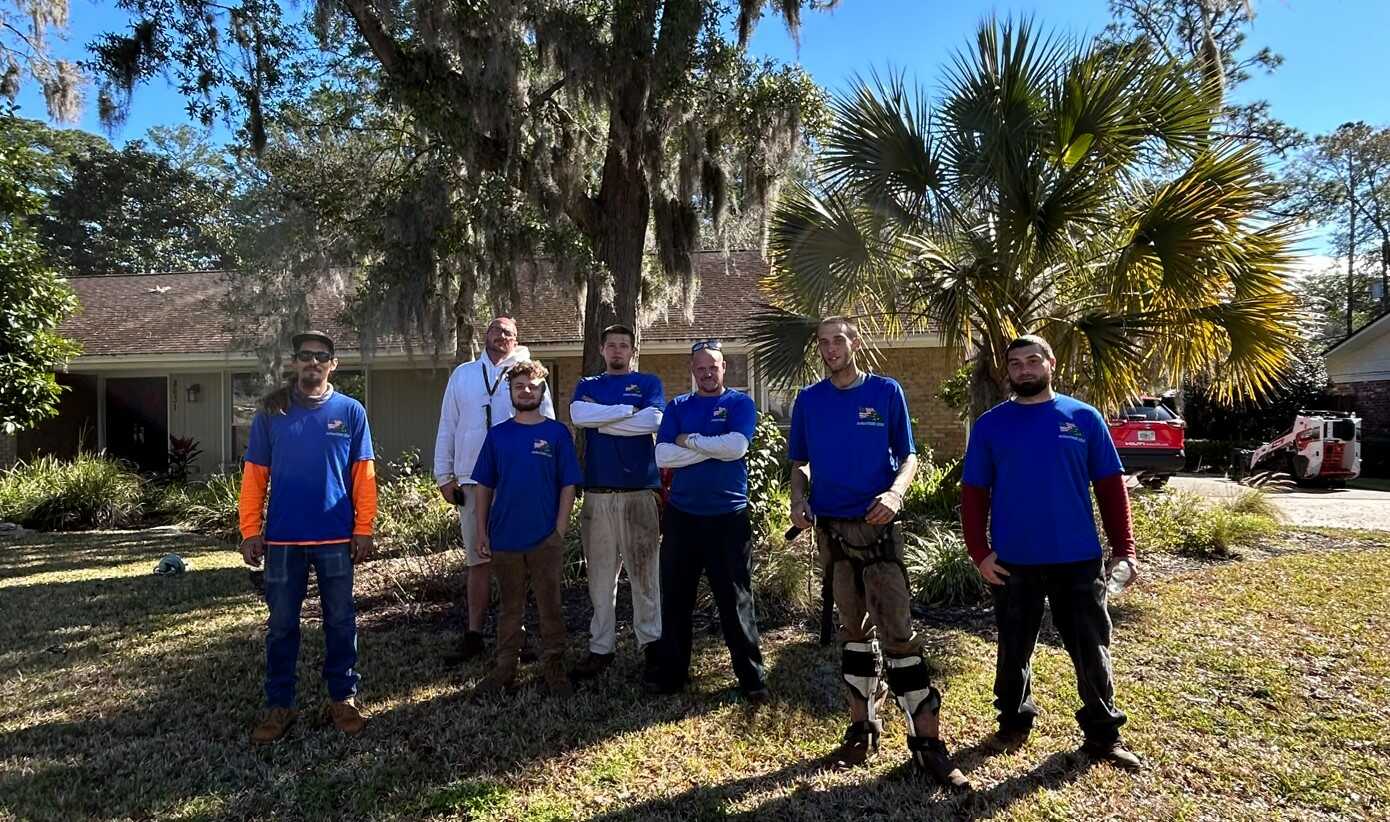 A team of seven tree service professionals posing in front of a house with lush greenery.